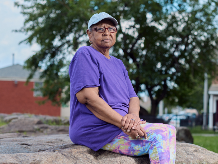A woman wearing an Oura Ring, abaseball cap, purple shirt, and colorful leggings sits on a rock and looks at the camera.