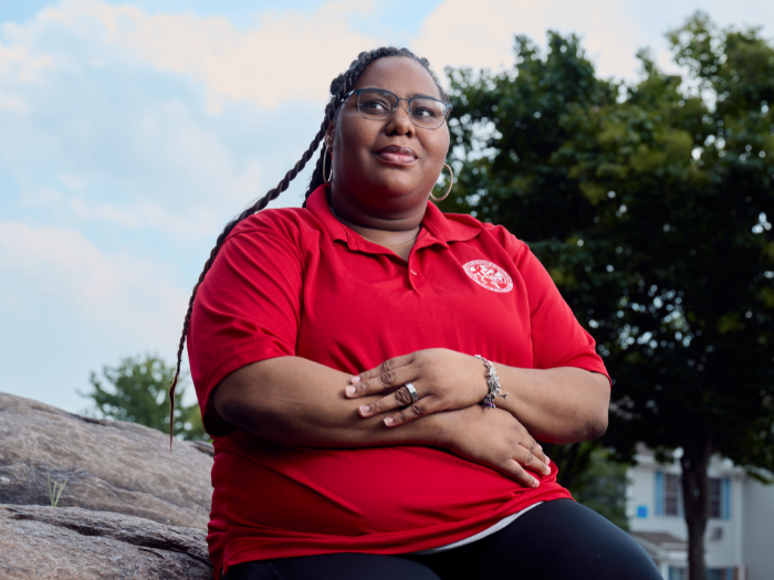 A woman wearing an Oura Ring, a red shirt, and glasses sits on a rock and looks off into the distance. 