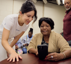 A woman wearing a white t-shirt with the OURA logo leans over a table to help another woman, wearing an Oura Ring, learn how to use the Oura App.